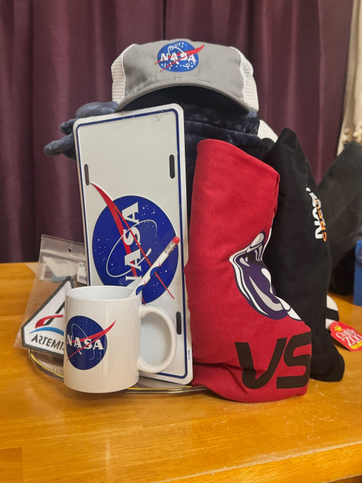 NASA-themed basket with cup, license plate, hat, blanket, 2 t-shirts, Artemis Mission patch, keychain, space shuttle stuffed toy, shot glass, and pencil, and a sticker
